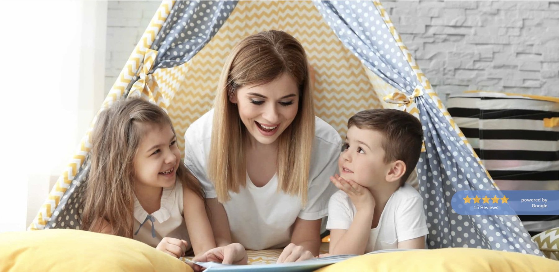A caregiver lying on the floor of a play tent reading a picture book with two young children.